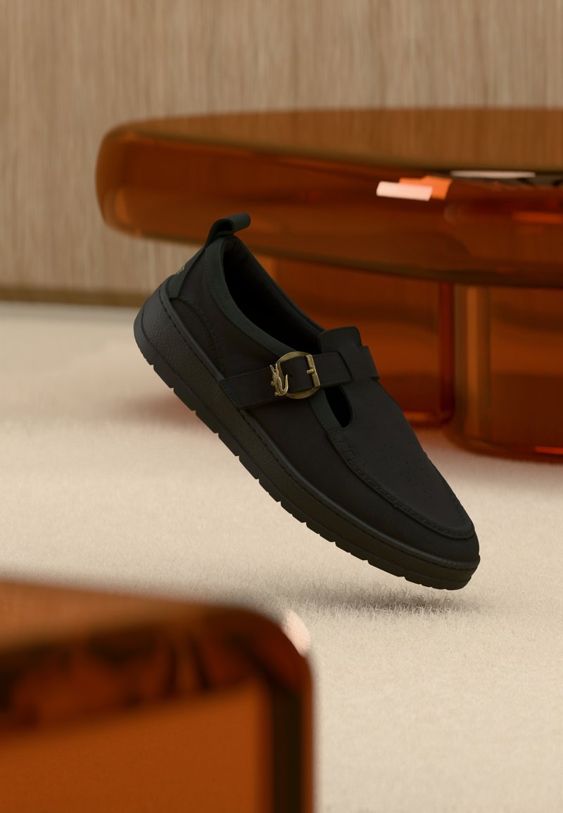 Scarpa nera slip-on con tomaia liscia, punta arrotondata e fibbia decorativa dorata. Presenta suole testurizzate e dettagli verdi.