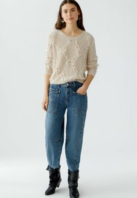 Beige gestrickter Pullover mit strukturiertem Rautenmuster, kombiniert mit hellblauen Baggy-Jeans. Das Modell trägt schwarze kniehohe Stiefel.