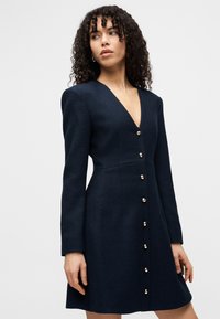 Γυναίκα με κατσαρά μαλλιά που φοράει ένα σκούρο μπλε φόρεμα V-λαιμόκοψη με μακριά μανίκια και μια σειρά από χρυσά κουμπιά στο μπροστινό μέρος, κοιτάζοντας στο πλάι.