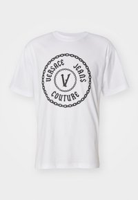 T-shirt en coton blanc avec un logo circulaire noir portant "VERSACE JEANS COUTURE" et un design central "V", entouré d'un motif de chaîne.