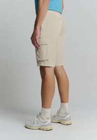 Beige cargoshorts met meerdere zijzakken, gemaakt van lichtgewicht stof. Gemaakt in combinatie met grijs en wit sportschoenen en beige sokken.
