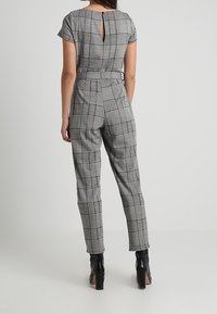 Šedý a černý kostkovaný overal s výřezem na zádech, krátkými rukávy, tapered nohavicemi a přiléhavým pasem. Nosený s černými vyššími botami.