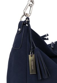 Coin d'un sac à main en cuir perforé bleu marine avec une fermeture à agrafe argentée, un gland en cuir et une étiquette métallique rectangulaire gravée de "SURI FREY".