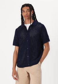 Camisa de punto de manga corta en color navy con cierre de botones en la parte delantera, que presenta tejidos abiertos y un cuello clásico. Textura suave y corte relajado.
