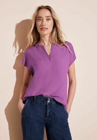 Blusa corta viola con scollatura a V, realizzata in un materiale morbido, abbinata a jeans in denim scuri.