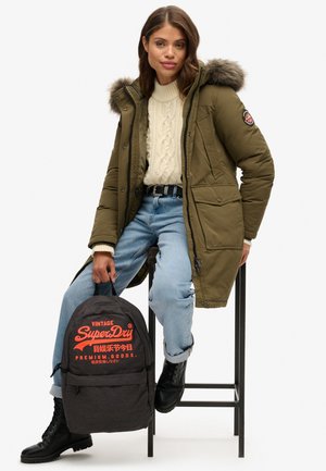 Parka vert olive avec une capuche doublée de fourrure, un pull en maille torsadée beige, un jean bleu et des bottes de combat noires, tenant un sac à dos Superdry noir.