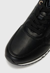 Scarpa sportiva nera con tomaia in pelle liscia, texture perforata, punta rotonda e suola piatta in gomma bianca con battistrada nero.