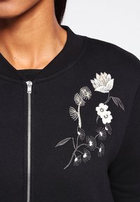 Sort sweatshirt med lynlås med hvid og cremefarvet blomsterbroderi langs halsudskæringen. Glat tekstur med ribbet krave.