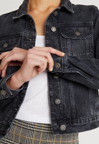 Zwarte spijkerjas met knoopdetails, twee borstzakken en een licht cropped pasvorm. Gedragen over een wit shirt, gecombineerd met een patroonrok.