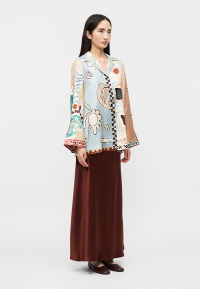 Γυναίκα που στέκεται φορώντας μακριά καφέ φούστα και φαρδιά, μακρυμάνικη πολύχρωμη μπλούζα με αφηρημένα σχέδια και μυτερό γιακά.