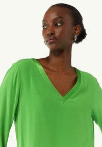 Green V-neck top made of smooth fabric, featuring a subtle sheen, relaxed fit, and accentuated neckline with stitching details.