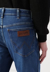 Niebieskie dżinsy z denimu z brązową skórzaną naszywką z napisem "Wrangler" na tylnej kieszeni, z pomarańczowymi przeszyciami i klasycznym wzornictwem.
