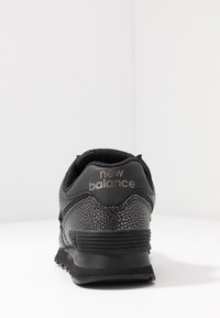 Schwarze Sneaker aus strukturiertem Material, mit einer glatten Ferse und reflektierenden Akzenten. "New Balance"-Logo auf der Rückseite angebracht.