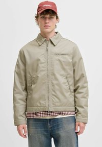 Ung mand iført en beige zip-up jakke, rød ternet skjorte under, blå jeans og en rød kasket med teksten "Originals".