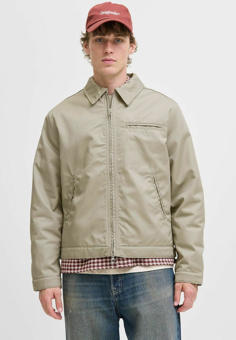 Ung mand iført en beige zip-up jakke, rød ternet skjorte under, blå jeans og en rød kasket med teksten "Originals".