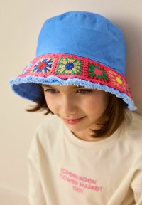 BlauerBucket Hat mit einem bunten gehäkelten Blumenband und fransigem Rand. Tragen von einem Kind mit dunklem Haar und einem hellfarbenen T-Shirt.