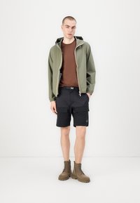 Chaqueta con capucha verde oliva y cremallera, camiseta marrón, pantalones cortos cargo negros y botas marrones con cordones. Bolsillos destacados en los pantalones cortos.