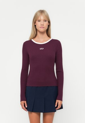 Long-sleeve, ribbed top in deep burgundy with a white crew neckline and "GAP" logo centered on the chest; paired with a navy pleated skirt.