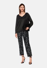 Frau in einem schwarzen Langarmoberteil mit weitem Kragen, glitzernden schwarzen Hosen, schwarzen High-Heel-Sandalen, die eine schwarze Feder-Clutch hält.