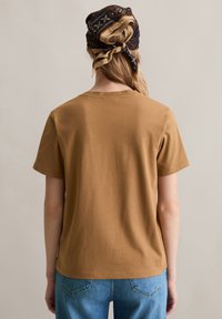 Kurze Ärmel, Rundhals-T-Shirt aus braunem Baumwollstoff, von hinten präsentiert. Kombiniert mit blauen Jeans.