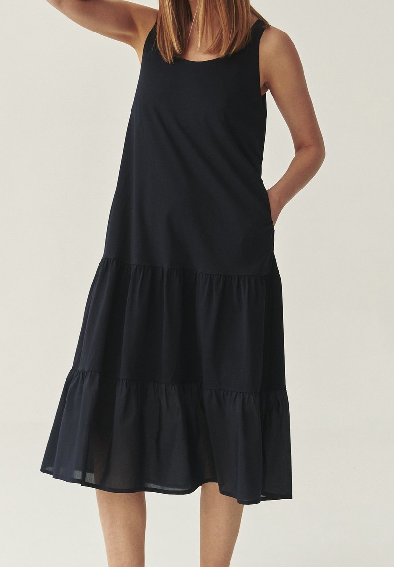 Femme portant une robe midi noire sans manches à volants avec un col rond, une main dans la poche, sur un fond clair uni.