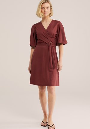 Frau trägt ein knieumspielendes, burgunderfarbenes Wickelkleid mit gepufften halblangen Ärmeln und einem Taillengürtel, kombiniert mit schwarzen Flip-Flop-Sandalen.