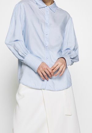 Camisa de botones azul claro con rayas, de mangas largas con puños fruncidos y un cuello clásico. Combinada con una falda blanca.
