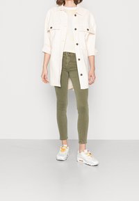 Camisa chaqueta blanca oversize con botones marrones, pantalones ajustados verde oliva y zapatillas blancas con detalles naranja.