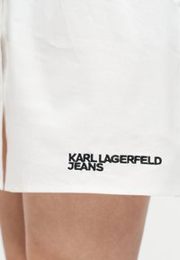Λευκό ύφασμα σε κοντινό πλάνο με μαύρη κεντημένη επιγραφή "KARL LAGERFELD JEANS" κοντά στο τελείωμα πάνω σε γυμνό δέρμα.