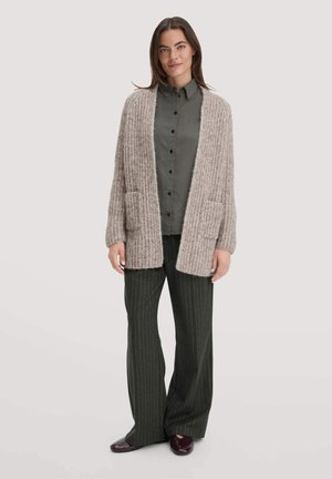 Beige gestrickte Strickjacke mit einem strukturierten Muster, offene Vorderseite und zwei vorderen Taschen, getragen über einem dunkelgrünen Hemd mit Knöpfen und gestreiften Hosen.