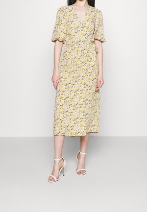 Femme portant une robe portefeuille jaune à fleurs avec des manches ballon et des escarpins blancs à bretelles à la cheville, debout devant un fond uni.