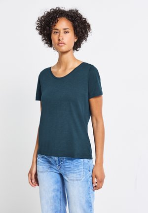 Mörk teal kortärmad t-shirt med rund halsringning, tillverkad av mjukt tyg, något avslappnad passform, i kombination med ljusblå jeans.