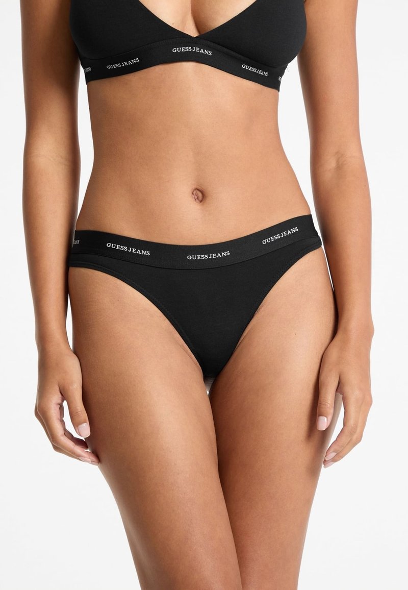 Black cotton thong underwear with a wide elastic waistband featuring repeating "GUESS JEANS" lettering in white. Smooth texture and minimal design.