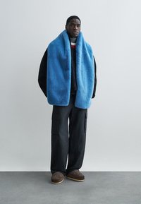 Μπλε εξωτερικό ένδυμα από ψεύτικη γούνα, μαύρο υφασμάτινο εσωτερικό στρώμα με υφή, σκούροι παντελόνια cargo και καφέ παντόφλες με λεπτομέρειες. Μινιμαλιστικό περιβάλλον.