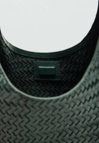 Interior de un bolso de cuero tejido verde, con un borde de textura suave y una pequeña etiqueta rectangular de cuero con costura.