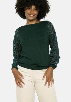 Pull vert foncé avec des manches en dentelle texturée et un col rond. Il présente des poignets ajustés et une coupe décontractée, associé à un pantalon de couleur claire.