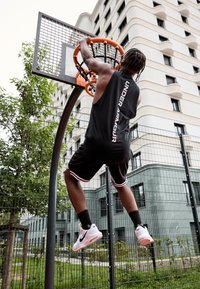 Maillot et short de basketball noirs avec des accents blancs, suspendus à un panier de basketball orange. Chaussures de sport blanches avec semelles roses. Végétation et bâtiments en arrière-plan.