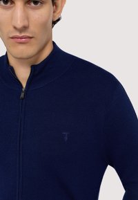 Marinblå zip-up tröja med högkrage, ribbat mönster och en liten logotyp på vänster bröst. Tyget ser stickat och mjukt ut.