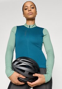 Garas piedurknes velosipēdu krekls teal un piparmētru zaļā krāsā, ar rāvējslēdzēja priekšpusi un ergonomisku dizainu; modelis tur melnu velosipēdistu ķiveri.