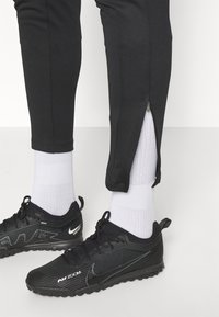 Pantalones deportivos negros hechos de un tejido texturizado, con cremalleras en los tobillos. Combinados con zapatillas Nike negras y calcetines blancos cortos.