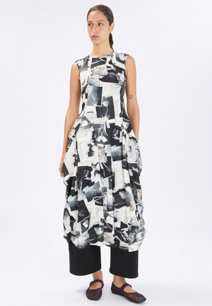 Donna con capelli raccolti che indossa un vestito senza maniche con stampa astratta in bianco e nero sopra pantaloni neri a gamba larga e ballerine scure.