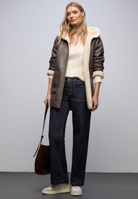 Ruskea shearling-takki, jossa on vaalea vuori, valkoinen v-pääntie-neule, tummat farkut ja vaaleat lenkkarit, asusteena ruskea olkalaukku.