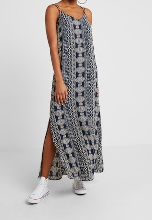 Femme portant une robe maxi sans manches à motif bleu marine et blanc avec des fentes latérales et des baskets montantes blanches sur un fond uni.