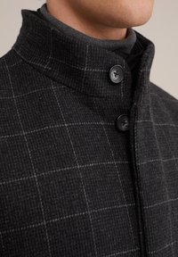 Mörkgrå ullkappa med hög krage, prydd med ett subtilt rutig mönster, två svarta knappar och en texturerad yta.