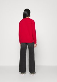 Felpa rossa con maniche raglan, texture liscia, polsini e orlo a coste; abbinata a pantaloni larghi grigi scuri e sneakers bianche.