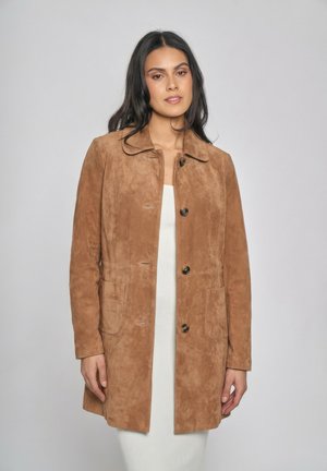 Donna con capelli lunghi e scuri che indossa un cappotto marrone chiaro in camoscio con bottoni sopra un vestito bianco a costine, in piedi contro uno sfondo semplice grigio chiaro.