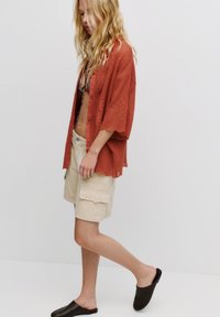 Rostfärgad, texturerad överdimensionerad knapp-up skjorta över en mönstrad bikini, i kombination med beige cargo-shorts och mörka slip-on skor.