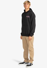 Sudadera negra con bolsillo frontal y logotipo bordado, combinada con pantalones caqui y zapatillas de skate negras con cordones blancos.