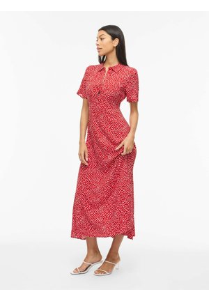 Mujer de pie, vistiendo un vestido midi rojo con lunares blancos y mangas cortas, combinado con sandalias blancas de tacón con tiras.