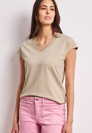 Kobieta ubrana w beżową bluzkę z krótkim rękawem i dekoltem w serek oraz jasnoróżowe dżinsy, stoi na tle jednolitego jasnego tła, patrząc lekko w górę i na prawo.